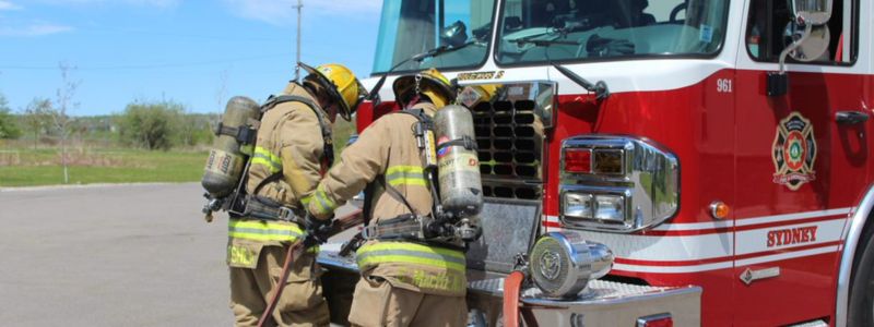 Two firefighters in full gear by a fire truck, seen from behind.